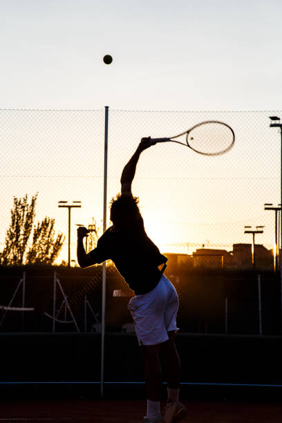 Back view of sportsman with tennis rocket hitting ball in back light on court.