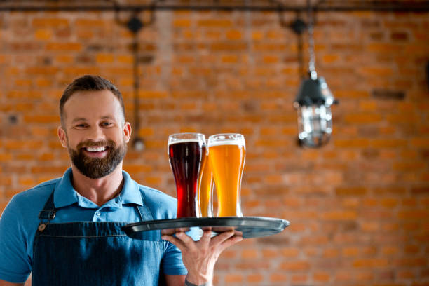 Happy bartender holding serving tray with glasses of beer Happy bearded bartender holding serving tray with glasses of beer, standing in the pub against brick wall. microbrewery photos stock pictures, royalty-free photos & images