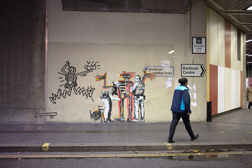 Woman walking past a Banksy mural in Golden Lane, near the Barbican Art centre in London. The famously anonymous street artists homage to the late New York grafitti artist, Jean-Michel Basquiat on walls near the Barbican Exhibition centre in London. It depicts a figure being frisked by two police officers, and is captioned with 