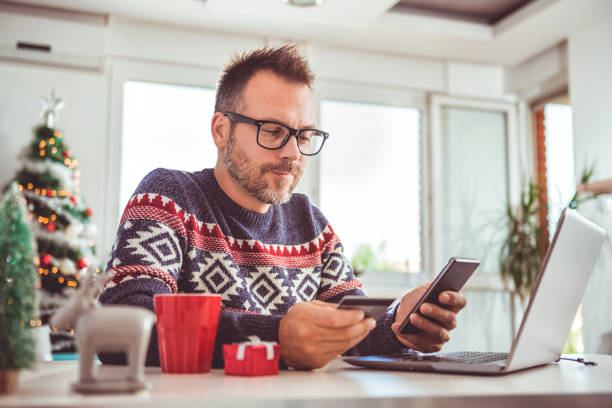 Men holding credit card and using laptop at home office Men wearing blue sweater and eyeglasses holding credit card and using laptop at home office during christmas holidays christmas sweater photos stock pictures, royalty-free photos & images