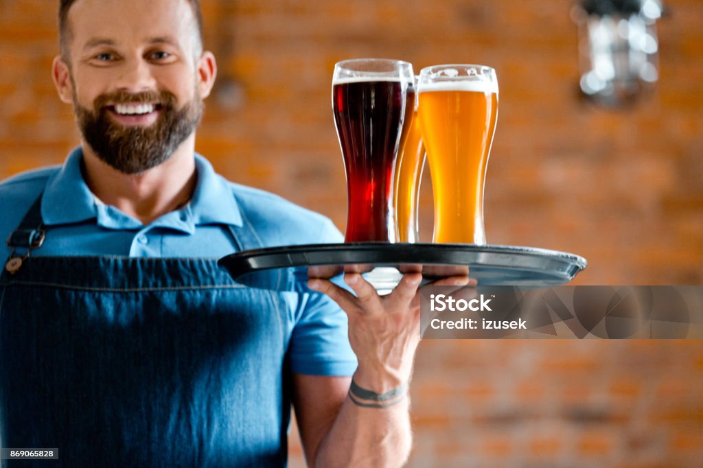 Happy bartender holding serving tray with glasses of beer Happy bearded bartender holding serving tray with glasses of beer, standing in the pub against brick wall. Tray Stock Photo
