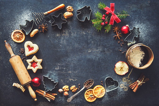 Christmas baking sweet food background with homemade cookies, spices, kitchen utensils, fir branches and red holiday decoration on dark rustic baking tray. Top view