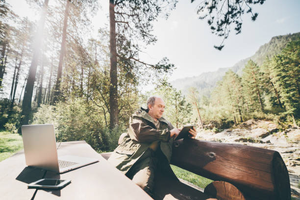 Elderly plus size man in forest with digital pad Mature forest warden is sitting on huge wooden bench with his gadgets and sending report using digital tablet; aged man in camping suit is having work session during his vocations in Altai mountains woodward stock pictures, royalty-free photos & images