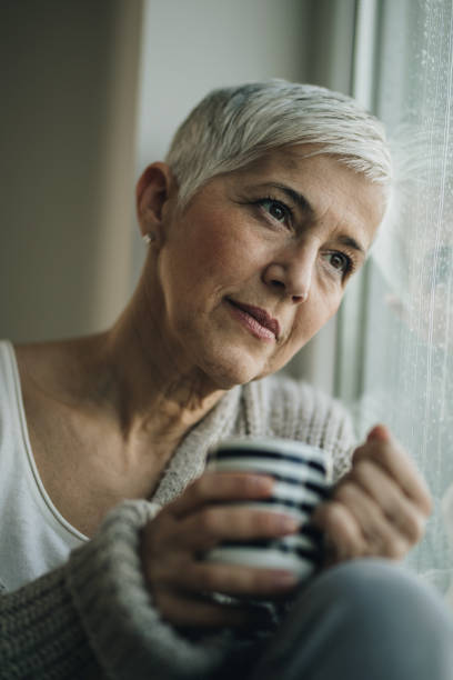 istock Thoughtful senior woman with tea cup looking through window during rainy day. 869245902
