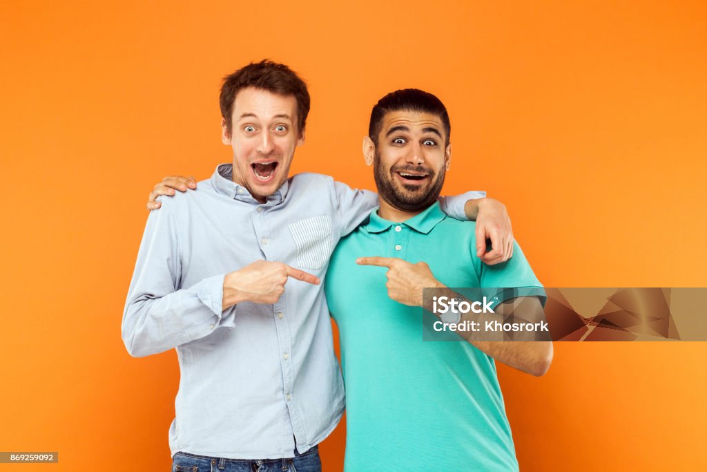 Two best friends hugging, pointing finger each other and looking at camera with shocked face Two best friends hugging, pointing finger each other and looking at camera with shocked face. Studio shot on orange background Friendship Stock Photo