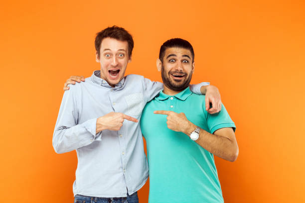 Two best friends hugging, pointing finger each other and looking at camera with shocked face Two best friends hugging, pointing finger each other and looking at camera with shocked face. Studio shot on orange background comedian photos stock pictures, royalty-free photos & images