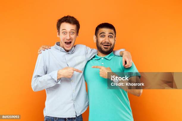 Two Best Friends Hugging Pointing Finger Each Other And Looking At Camera With Shocked Face Stock Photo - Download Image Now