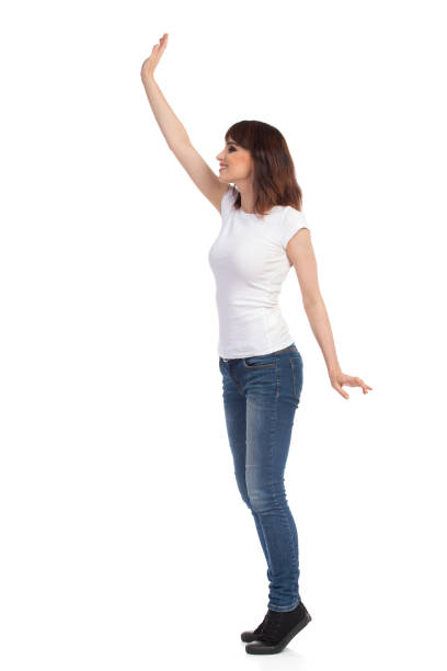 Young Woman In White T-shirt And Jeans Is  Standing TipToe And Waving Hand. Side View. Happy young woman in jeans and white t-shirt is standing on tiptoes, rising arm, waving, looking away and smiling. Full length studio shot isolated on white. tiptoe stock pictures, royalty-free photos & images