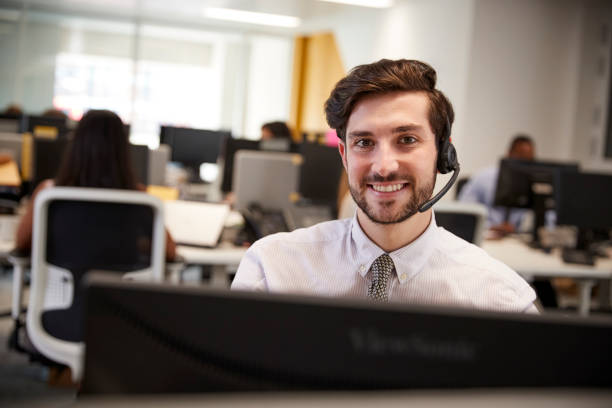 istock Young man working at computer with headset in busy office 869286458