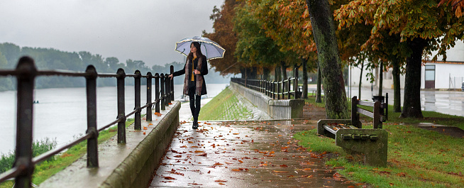 Beautiful young woman with umbrella walking alone beside the river on a rainy autumn day.