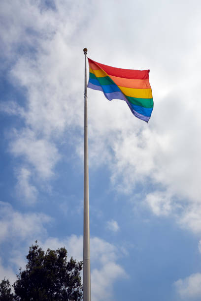 the Pride Flag on a tall pole against a blue skies and puffy white clouds stock photo