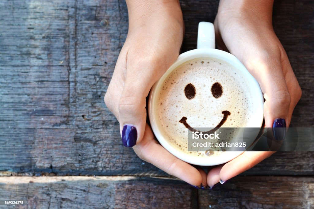 Cara sonriente en la espuma del capuchino, manos de mujer sosteniendo una taza de capuchino en mesa de madera - Foto de stock de Cara sonriente antropomórfica libre de derechos