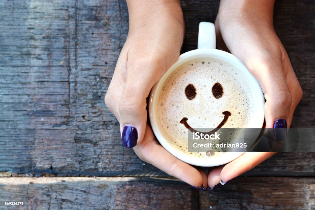 Smiley face on cappuccino foam, woman hands holding one cappuccino cup on wooden table Anthropomorphic Smiley Face Stock Photo