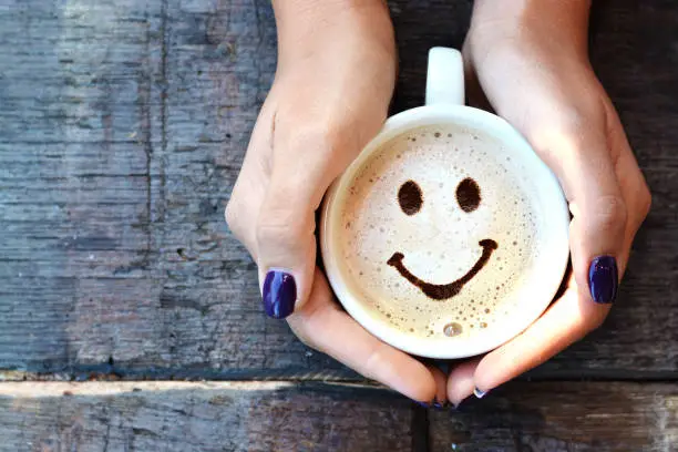 Photo of Smiley face on cappuccino foam, woman hands holding one cappuccino cup on wooden table