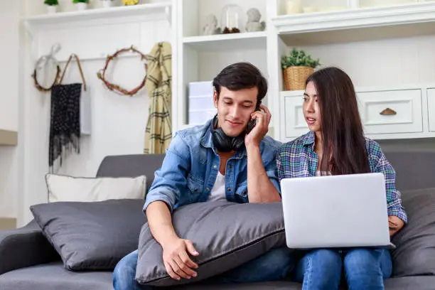 Young Asian couple relaxing together on sofa and using mobile-phones at home in the living room