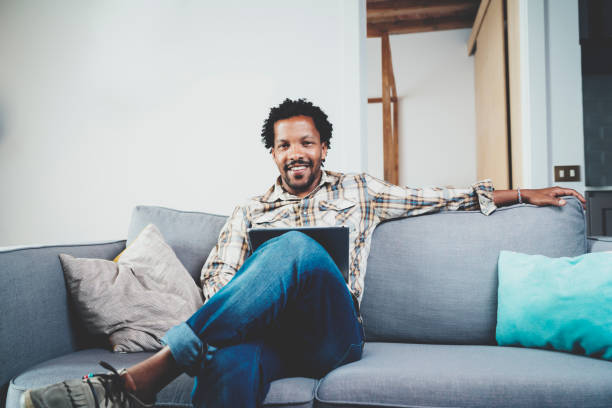 istock Bearded smiling American African man using tablet for video conversation while relaxing on sofa in modern home.Concept of young business people working at home.Blurred background.Front view. 869344990