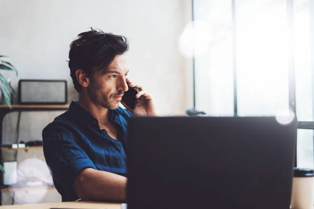 istock Handsome young businessman working at sunny work place on laptop.Elegant Man using smartphone while sitting at the wooden table.Blurred background.Horizontal. 869348802