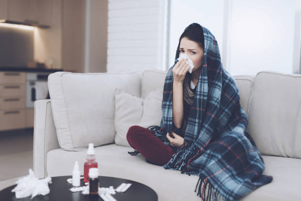 istock A cold woman sits on a light couch wrapped in a blue checkered whip. She blows her nose into a napkin 869356080