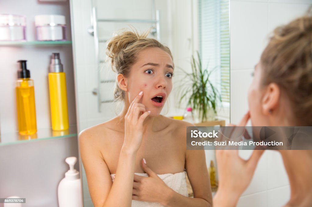 Young beautiful woman with pimple Young beautiful woman surprised to see pimple on skin. Unhappy girl with stunned funny face standing in towel in bathroom, looking in the mirror, pointing to zit Acne Stock Photo