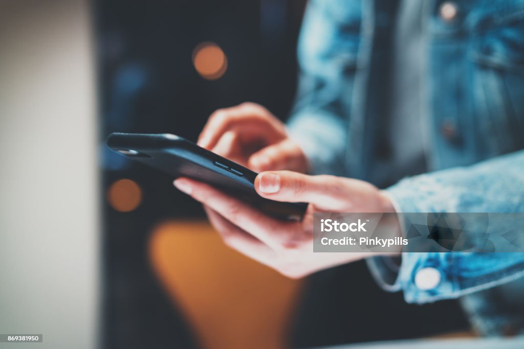 Vista de cerca de manos femeninas sosteniendo moderno teléfono inteligente y apuntando los dedos sobre la pantalla táctil por la noche. Fondo horizontal, borrosa, efectos bokeh. - Foto de stock de Teléfono móvil libre de derechos