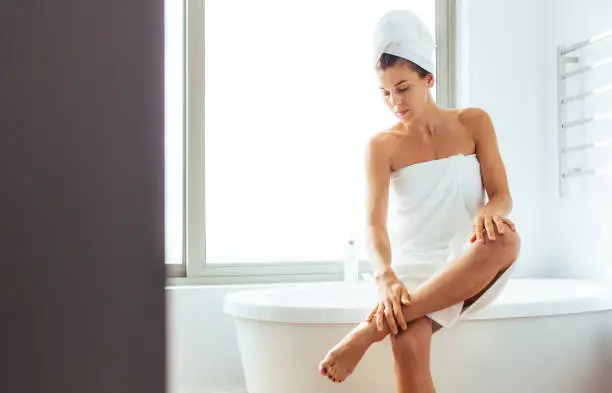 Woman applying moisturizer to her legs after bath. Woman in towels wrapped around head sitting on bathtub in bathroom.