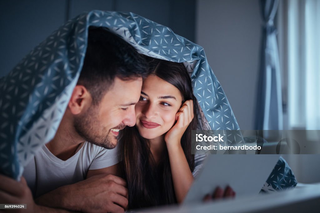 Their sleepless nights are filled with love A happy couple are lying in bed under covers while watching a romantic film on the tablet. Couple - Relationship Stock Photo