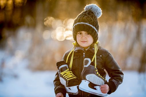 An Ethnic boy is outdoors on a sunny winter day. He is wearing a jacket and hat. He is standing on the snow and smiling at the camera while holding a pair of hockey skates.