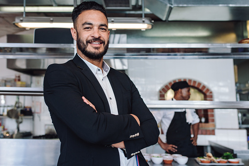 Portrait of young businessman standing in his restaurant with staff in kitchen. Proud restaurant owner standing with his arms crossed and looking at camera.