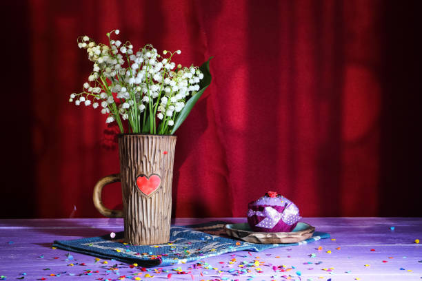 bouquet de fleurs de lys-de-la-vallée dans une tasse en céramique avec un coeur sur un cupcake de peluche bleue pour serviettes de papier et textile et confettis sur une table violette sur fond bordeaux - claret cup photos et images de collection