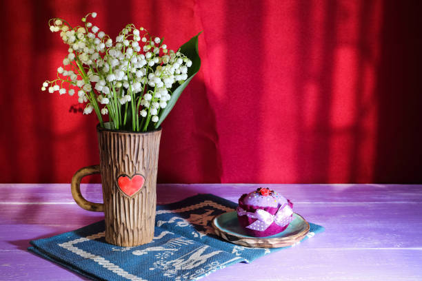 bouquet de fleurs de lys-de-la-vallée dans une tasse en céramique avec un coeur sur un bleu textile et serviette cupcake de peluche sur table violet sur fond bordeaux - claret cup photos et images de collection