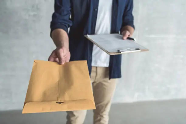 This is for you. Close-up of envelope in hand of diligent professional courier. He is standing with package and personal contract against gray wall background. Selective focus