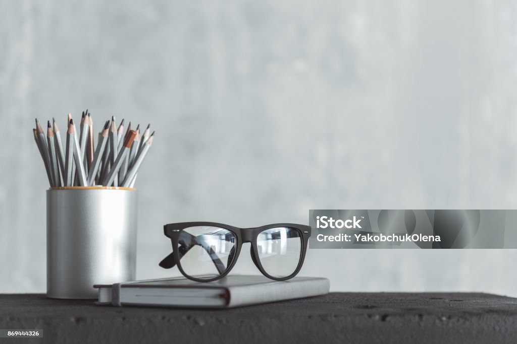 Personal things of skillful worker Prepared for work. Close-up of stylish eyeglasses of professional employee is on notepad and box of pencils is near. Gray wall in background. Copy space in the right side Desk Stock Photo