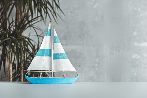 Vacation concept. Close-up of model of yacht is on table at modern office. Gray wall and houseplant in background. Copy space in the right side