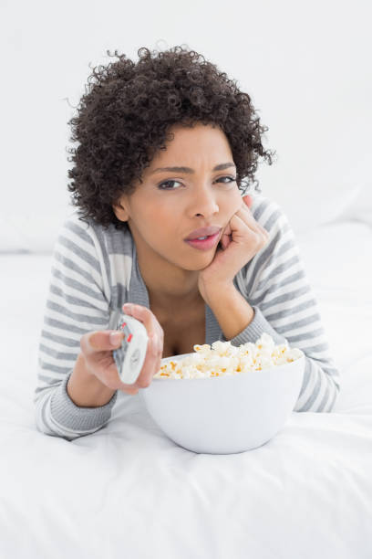 istock Woman with remote control and popcorn bowl lying in bed 869460020