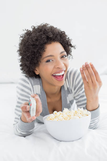 istock Cheerful woman with remote control and popcorn bowl in bed 869460068