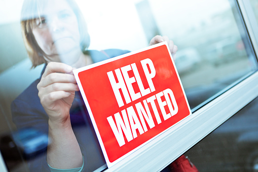 Store owner posting a HELP WANTED sign on her retail store window for hiring new employees. Employment opportunities available as economic recovery.