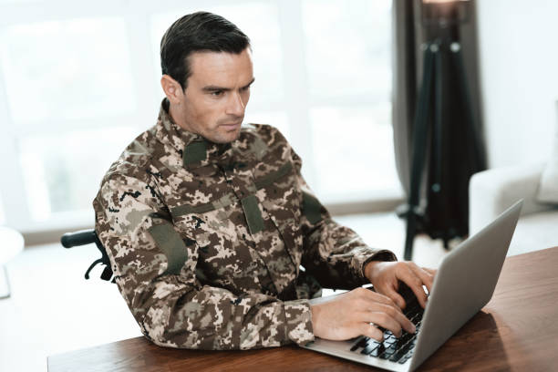 istock A disabled military man is sitting at a table and working with a laptop. Behind it is a large panoramic window. 869485710