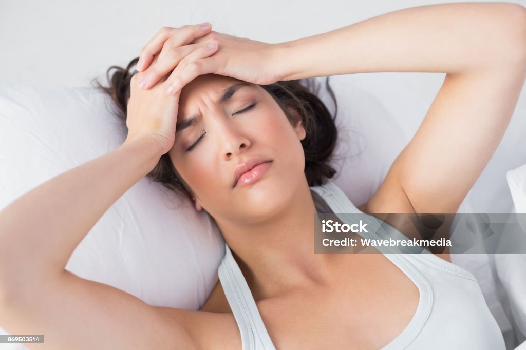 Sleepy woman suffering from headache in bed High angle view of a sleepy young woman suffering from headache with eyes closed in bed at home 20-24 Years Stock Photo