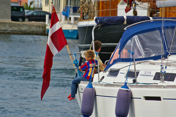 桑德堡, 丹麥-2012年7月5日-年輕男孩與足球襯衫坐在 gunwhale 的白色帆船遊艇與丹麥國旗旁邊, 他看著水 - 利安奴·美斯 個照片及圖片檔