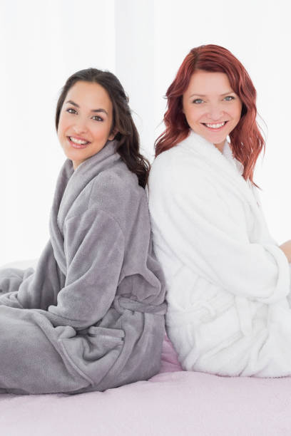 istock Female friends in bathrobes sitting back-to-back on bed 869507064