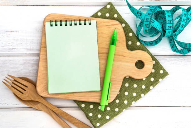 istock Notepad on chopping board with wooden fork and spoon and the measuring tape on white table , recipes food or diet plan for healthy habits shot note background concept 869558358
