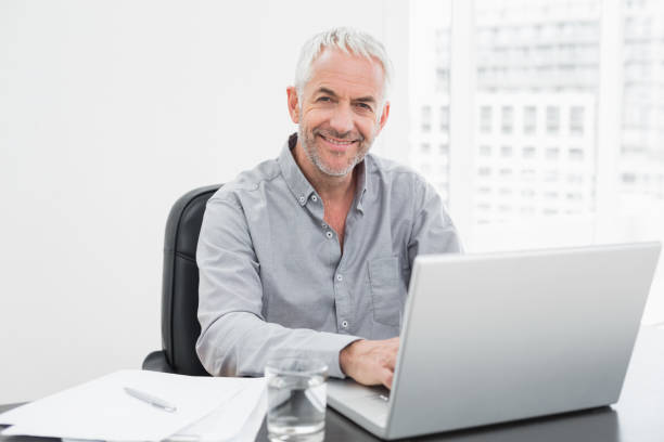 istock Smiling mature businessman using laptop at desk in office 869570370