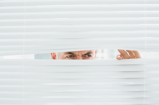 Closeup of a serious mature businessman peeking through blinds in the office