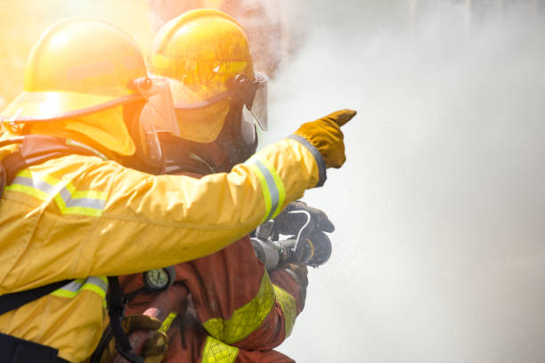 two firefighters water spray by high pressure nozzle to fire surround by smoke with flare and copy space stock photo