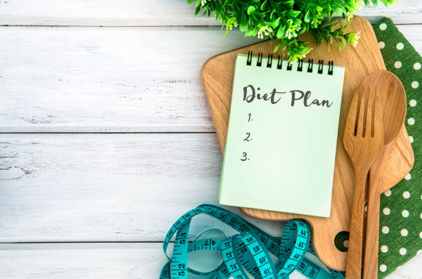 The Notepad with Diet Plan list text  on chopping board with wooden fork and spoon and measuring tape on white table , recipes food or diet plan for healthy habits shot note background concept