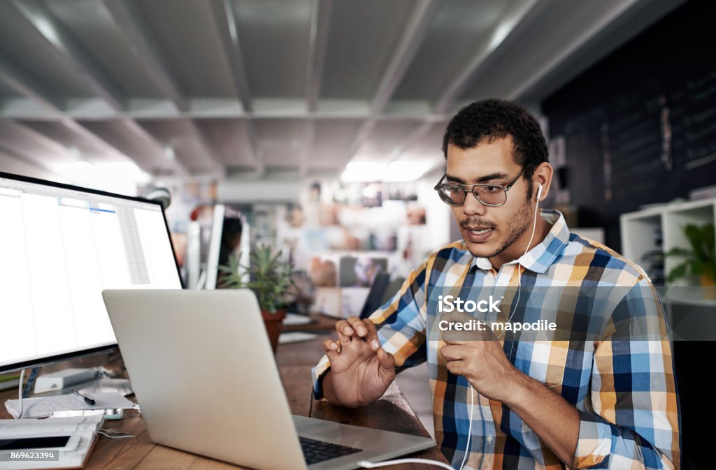 Giving virtual advice Shot of a young businessman using earphones and a laptop at his desk in a modern office Video Conference Stock Photo