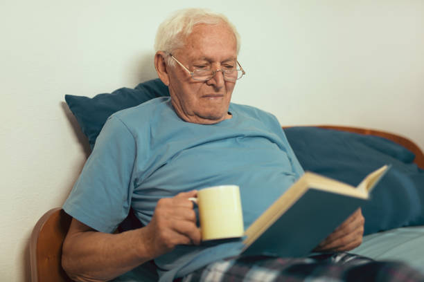 istock Senior man lying on bad and reading book at home 869645128
