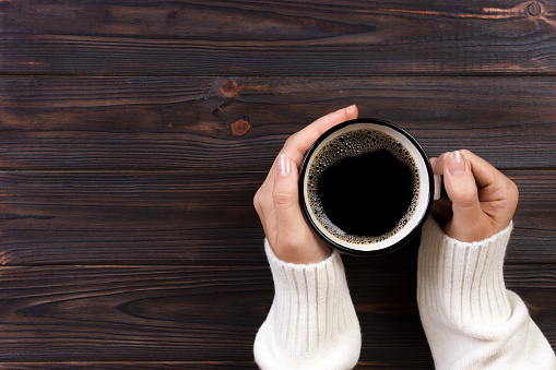 Lonely woman drinking coffee in the morning, top view of female hands holding cup of hot beverage on wooden desk.