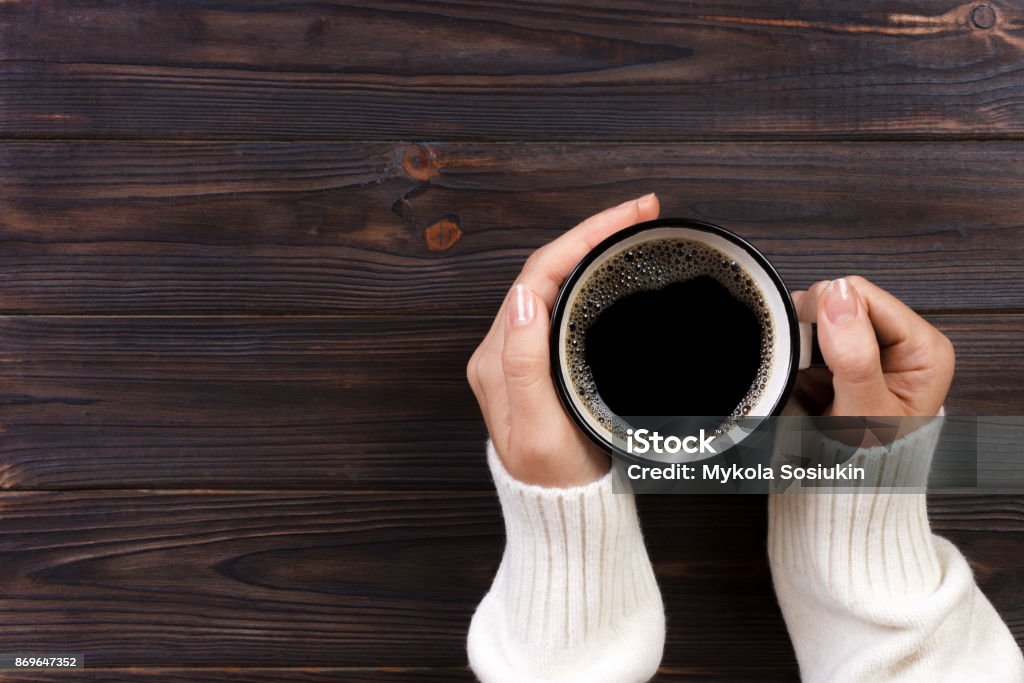 Lonely woman drinking coffee in the morning, top view of female hands holding cup of hot beverage on wooden desk Lonely woman drinking coffee in the morning, top view of female hands holding cup of hot beverage on wooden desk. Coffee - Drink Stock Photo