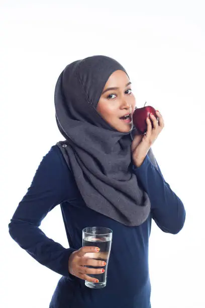 A young beautiful hijab sportsgirl is biting an apple with plain water looking at camera isolated with white background